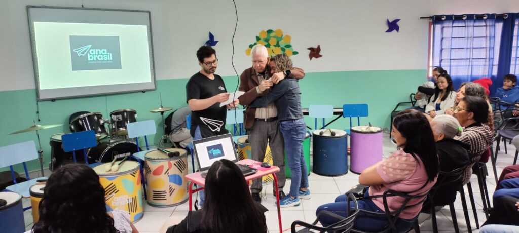 Participante idoso abraça uma educadora na sala de música do CCII, ao lado dos tambores do Batuque