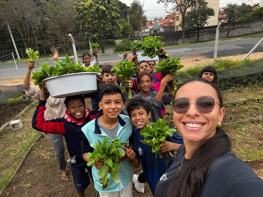 Crianças do ANA DIC sorriem mostrando as verduras que colheram na horta da unidade, ao lado da educadora